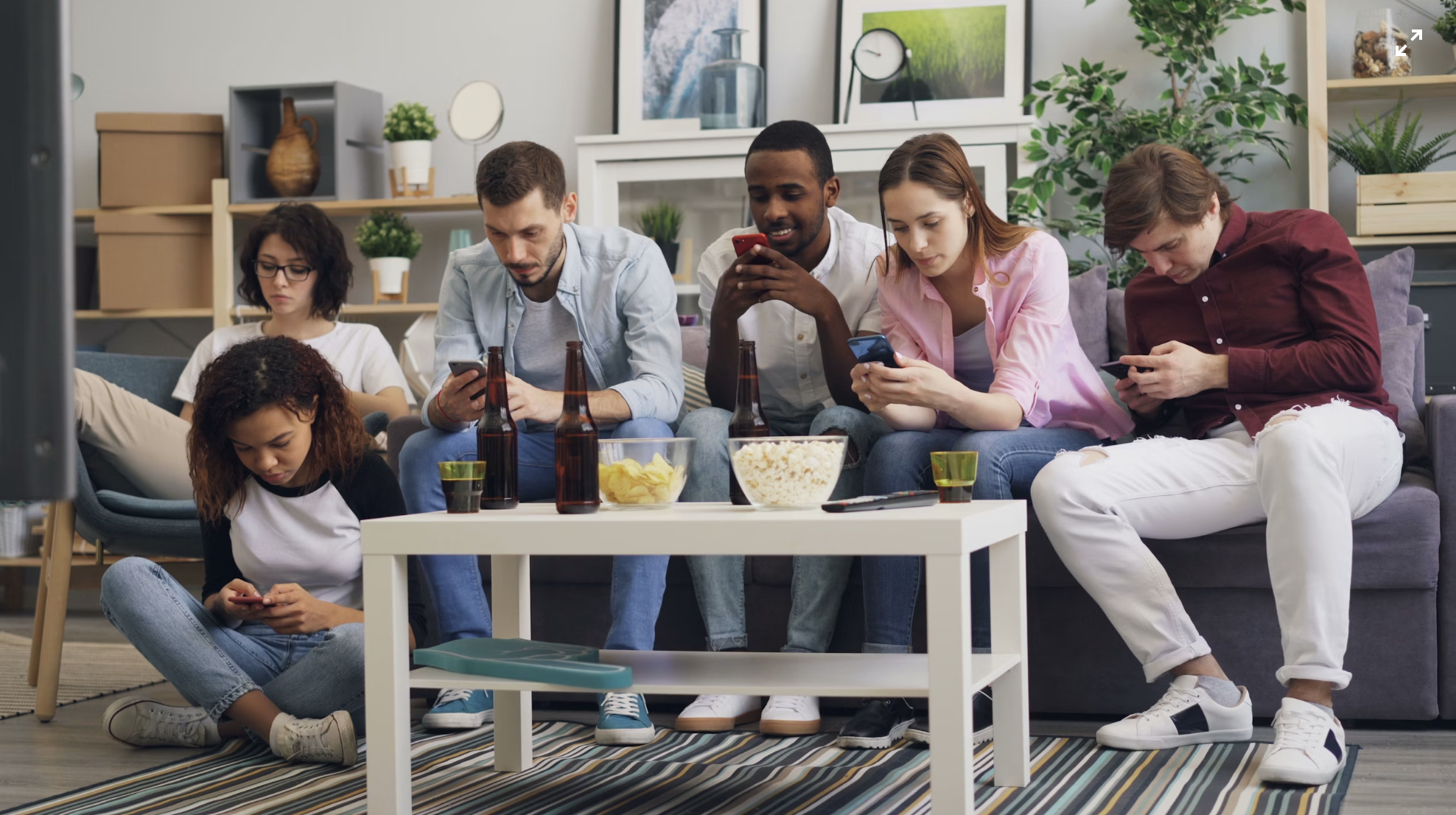 A group of diverse people sitting on a couch, each using their smartphones, with snacks and drinks on the table in front of them.